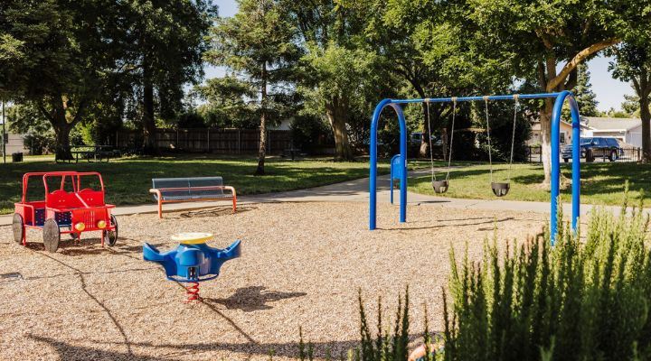 Image of a blue playground spinner, two bucket swings, a red car-climber, and a park bench