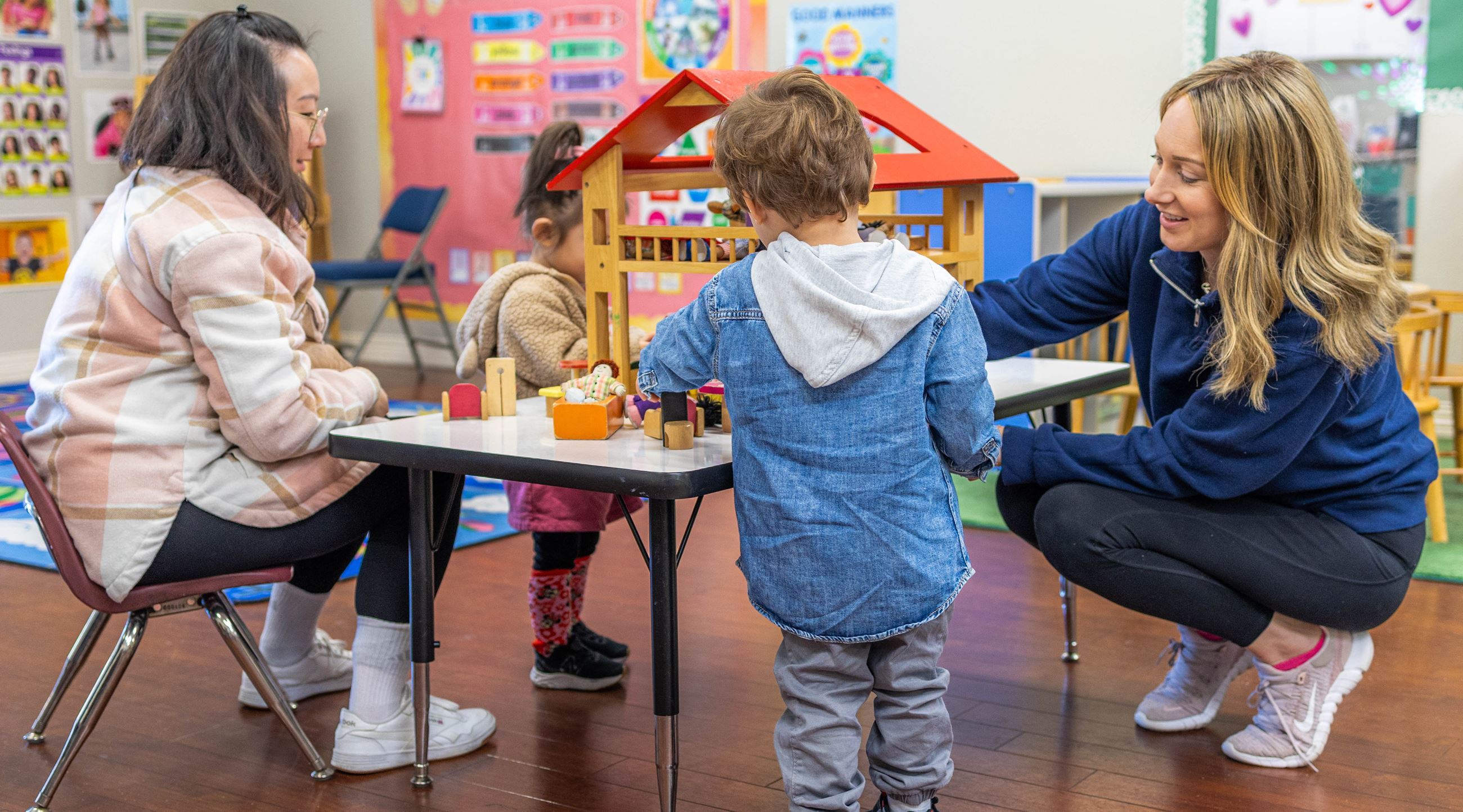 A parent, a teacher, and two toddlers gather around a dollhouse to play together.