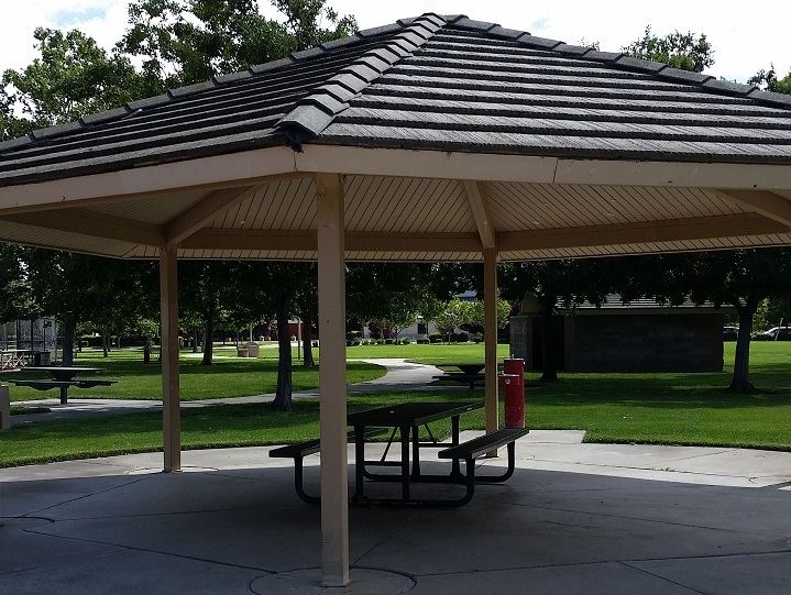 Tan colored covered picnic area with 1 metal table underneath.  Trees and grass in the back ground.